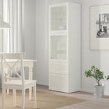 Modern white BESTÅ cabinet, tall with glass top & bottom shelves, solid body, sleek design, fits neatly in dining space.