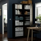 Black BESTÅ storage unit with adjustable shelves, holding books, boxes, and decorative items. Next to a wooden desk with a lamp and chair.