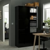 Black BESTÅ storage unit with adjustable shelves and push-open doors, filled with books and boxes, near a wooden desk with a chair and a window.