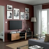 Modern living room with dark red walls, BESTÅ tv bench, mounted screen, shelves, black-and-white photos, glass coffee table, lamp, large window.