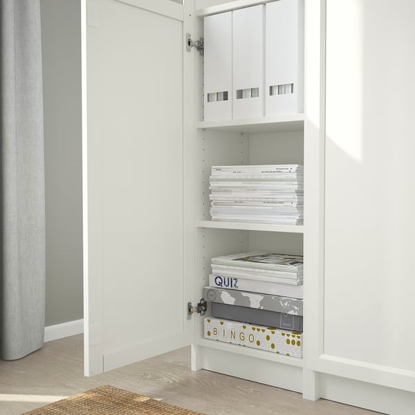 A white BILLY bookcase with adjustable shelves holding books and boxes, placed in a corner, anchored to the wall.