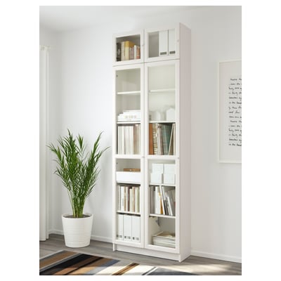 White BILLY bookcase with glass doors, showcasing books and decor, placed near a plant and wall art.