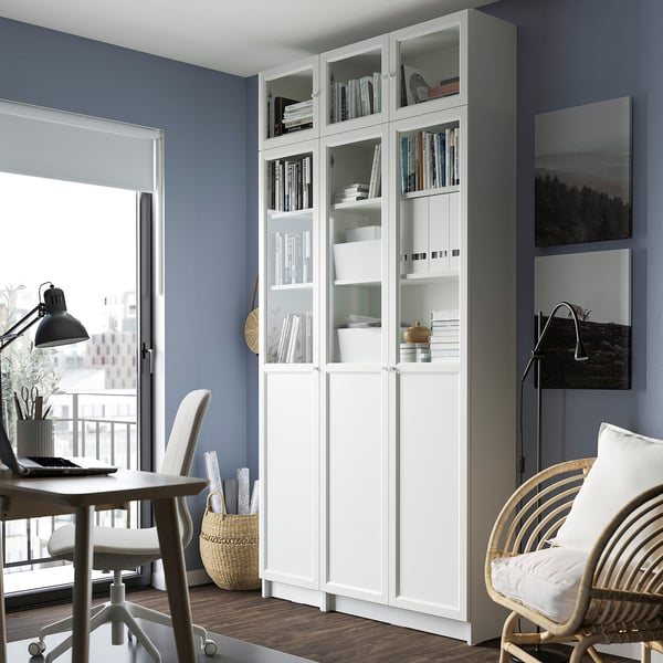 BILLY white bookcase with glass doors between desk and wicker chair, blue walls, books, storage, near window.