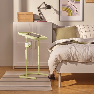 Small yellow-green BJÖRKÅSEN laptop stand next to a large bed, white cupboard, and a striped rug.