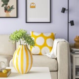 Bright yellow and white striped vase on table, holding green succulents. Nearbeige sofa with bold yellow polka dot pillow and wall frames.
