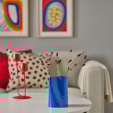 Blue vase with red flowers on table near beige chair, red candle, art on wall.