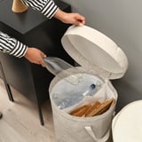 Person discarding a plastic bottle into an open DIMPA waste sorting bag. The bag is partially filled with recyclable items and has a lid.