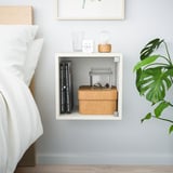 Small white EKET wall shelf holds books, boxes, and plants near bed.