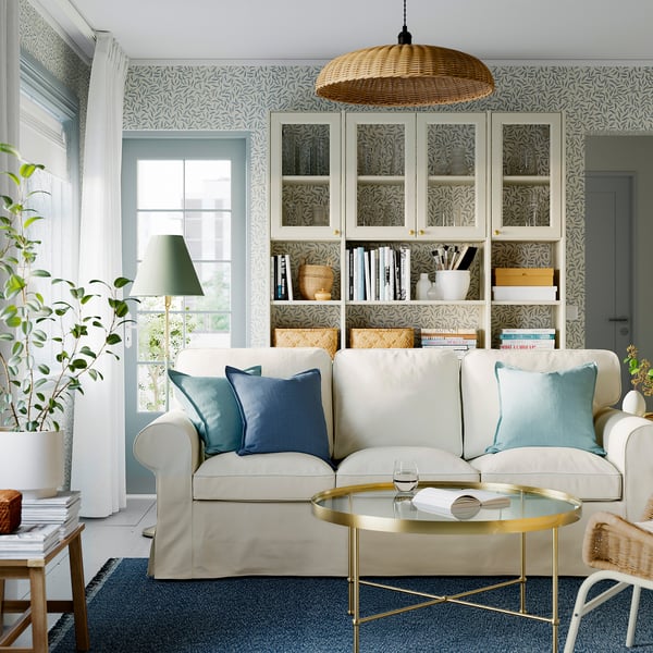 Modern living room with white EKTORP sofa, blue pillows, glass coffee table, patterned wallpaper, and bookshelf.