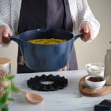 Person holding a blue pan with yellow food, resting on a decorative bamboo heat-resistant stand on a table with other ingredients.