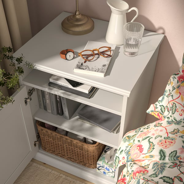 Bedside table with drawers, holding books, watch, glasses, vase, and lamp.