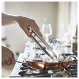 A person is cooking with FINMAT tweezers, gripping sizzling food in a copper pan on a stove, steam rising, stainless steel design.