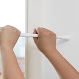 Hands installing a FJÄDRANDE curtain rod between window frames, showcasing its no-drill, spring mechanism.