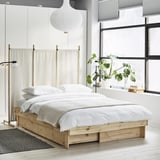 Modern minimalist bedroom with wooden extendable bed, white linens, large windows, and hanging plants.