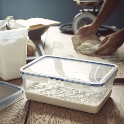 Clear plastic food container sits on wooden table, showing flour residue, with person handling dough in background.