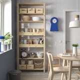 Wooden IVAR storage unit with kitchen items and blue clock in dining area.