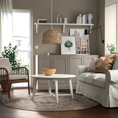 Cosy living room with JÄRNVÄG rug, pastel tones, coffee table, chair, cabinet, lamp, and wall decor; natural light from window with balcony view.