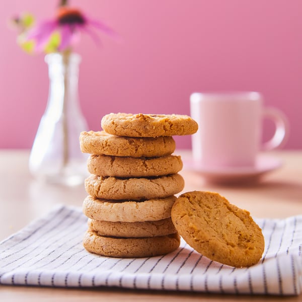 A stack of golden-brown KAFFEREP cookies on a striped cloth, with a white mug and vase with flowers in the background.