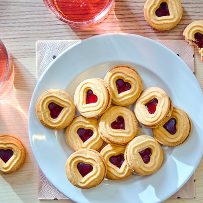 Plate of heart-shaped jam-filled cookies, golden-brown, with pinkish-red jelly centres, and glasses of pink liquid beside it.