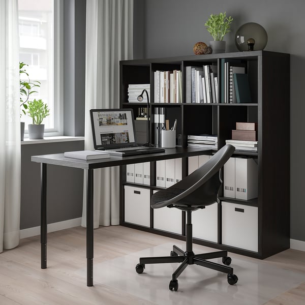 Modern black KALLAX shelf unit with table and chair, displaying books, plants, and a laptop, occupying a whole wall.