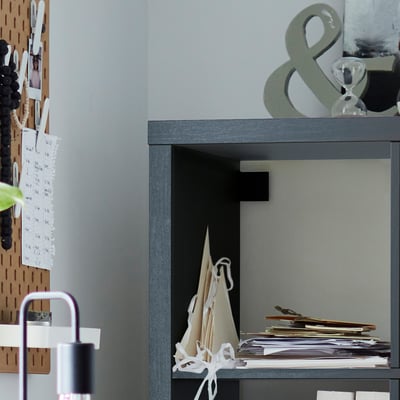 Modern grey KALLAX shelving unit with various items, including a lamp, plant, and papers, against a light wall with a cork board.