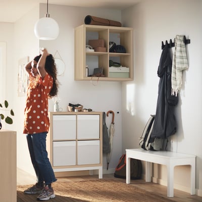 Person dancing near KALLAX shelf with jackets, bench, and plant in bright entryway.