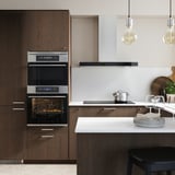 Kitchen with dark wood cabinets, ovens, hanging lights, white counter, and cooking setup.