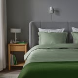 Tidy bedroom with grey bed, green sheets, LÄKEVÄNDEROT bedding, bedside table with lamp, wine glass, books.