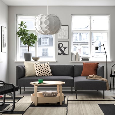 Modern living room: grey LILLEHEM sofa with cushions, wooden coffee table, large window, black chairs, green plant, and unique light fixture.