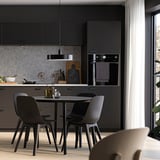 Modern kitchen with black METOD cabinets, round dining table, and grey chairs. Large window with white curtains. Black oven and gold vase.