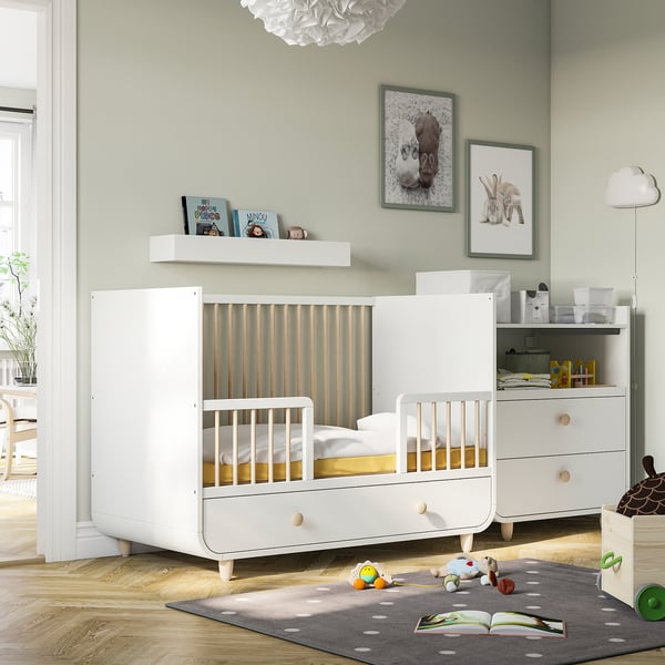 A white MYLLRA cot with drawer, adjustable sides, in a nursery filled with books, a dresser, and toys.