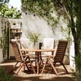 Outdoor dining set with acacia wood table and chairs. White cushions, string lights above, greenery around.