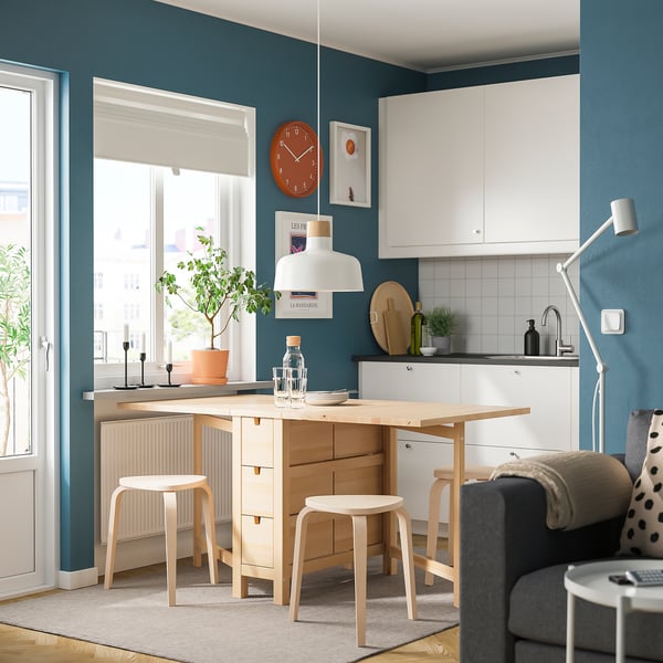 Modern kitchen with light wood table & stools, white cabinets, grey walls & large window.