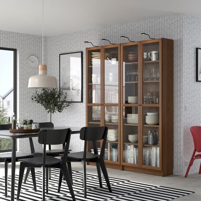 Modern dining room with a tall wooden OXBERG bookcase, glass doors, dishes, and a black dining set. Large plant and striped rug.