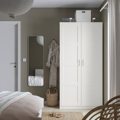 Minimalist bedroom with white PAX wardrobe, mirror, basket, and soft-close hinge. Bed in foreground with wicker chair.
