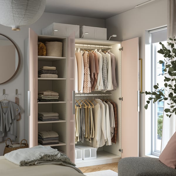 A pastel pink wardrobe with clothes hangers and shelves, featuring interior lighting, neatly organised clothes, and storage baskets.