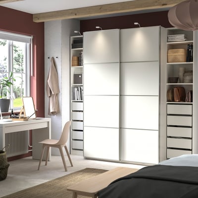 Modern bedroom with white PAX wardrobe, sliding doors, and desk area. Light wood accents and beige chair.