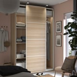 Modern bedroom with light wood PAX wardrobe, pink walls, hanging clothes, folded items, and decorative pieces.