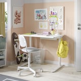 White kids desk with wheels, featuring a bright yellow backpack, art prints, and various stationery items on a light wooden floor in a cosy room.