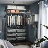 A tidy closet with clothes, shelves, and baskets. Dark blue walls with white furniture, featuring a round mirror and a small plant.
