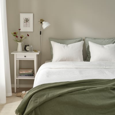 Minimalist bedroom: white bed with green blanket, bedside table, lamp, books, vase with flowers.
