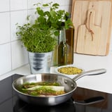 Metal pan cooking fish with herbs beside green plant and wooden board on stovetop.