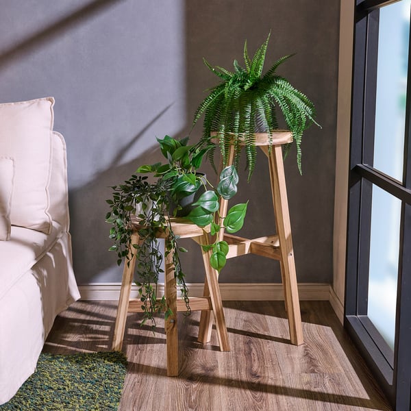 SKOGSTA brown wooden plant stand holds large greenery near white bed, fits corner space.