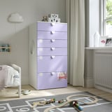 Lavender SMÅSTAD kids dresser with 6 drawers, fitting neatly in a corner beside a white crib and near a window with light-colored curtains.