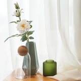 Clear vase holds white flowers with green leaves, next to two decorative vases on a wooden surface by white curtains.