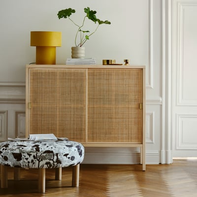 Rattan stockholm cabinet with brass handles, lamp, vase, and ottoman.