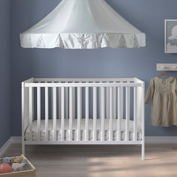White SUNDVIK baby crib with canopy against blue wall, beige dress nearby.