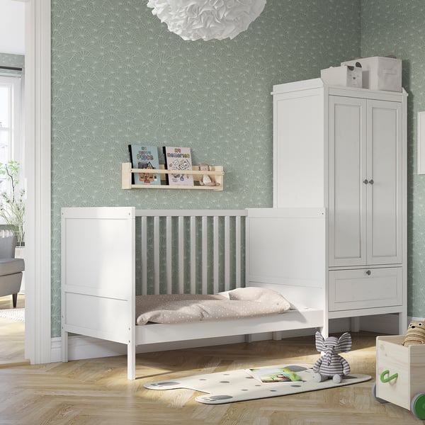 Small white SUNDVIK baby cot & changing unit in modern nursery with wall shelf & toys.