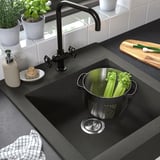 Black sink with vegetables in strainer, soap dispenser, and kitchen plants.