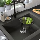 Black farmhouse sink with stainless colander, holding celery. Dark quartz, deep basin for large items.